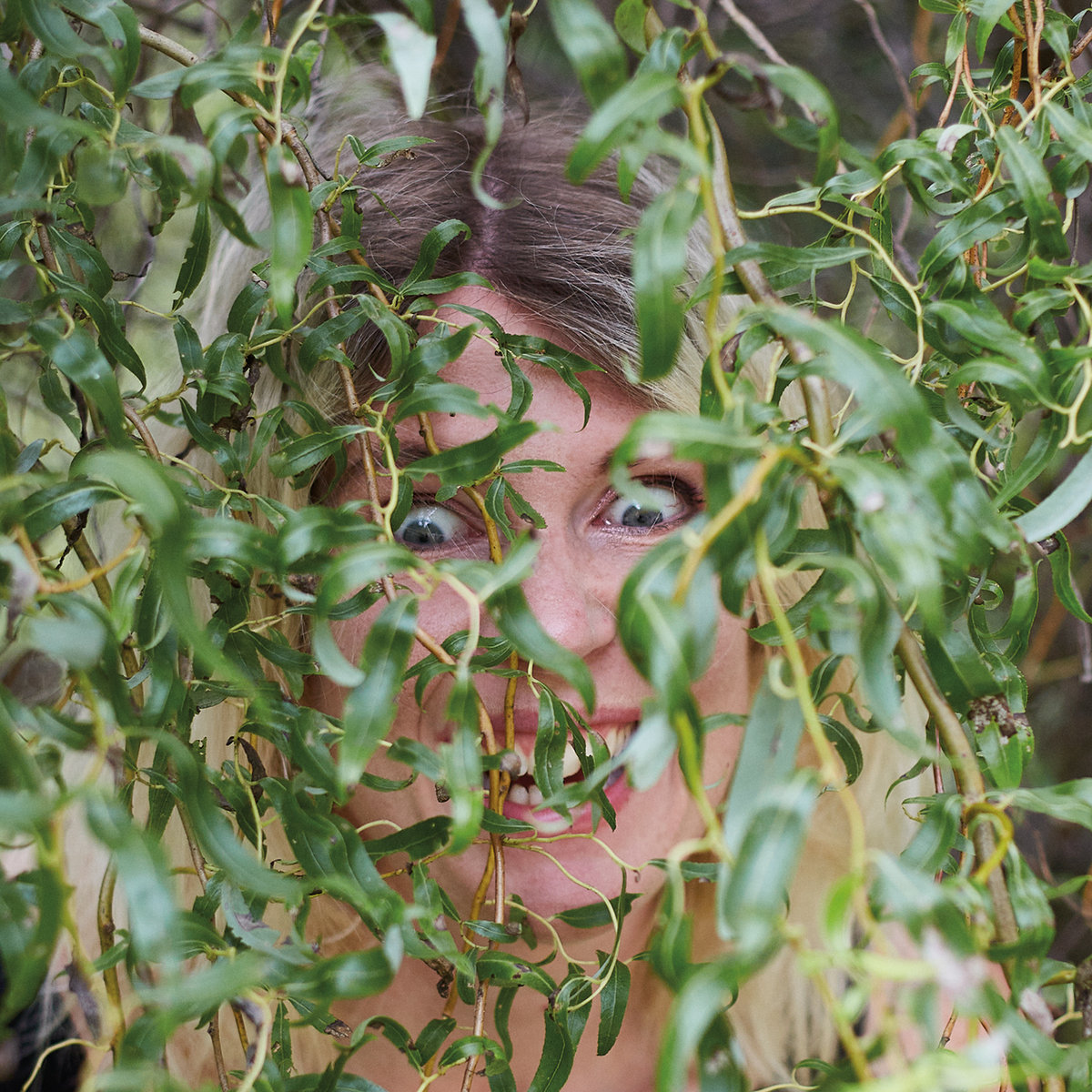 Bite Down album cover. Rosali's face with wide eyes and smile hidden behind the thick leaves and branches of a plant.